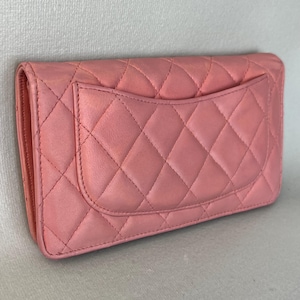 May include: A pink quilted leather wallet with a flap closure. The wallet features a diamond pattern and a small pocket on the back. The color is a soft, muted pink.