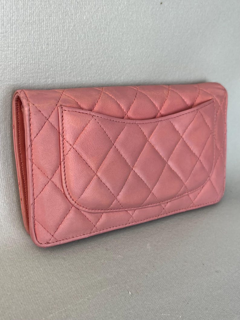 May include: A pink quilted leather wallet with a flap closure. The wallet features a diamond pattern and a small pocket on the back. The color is a soft, muted pink.
