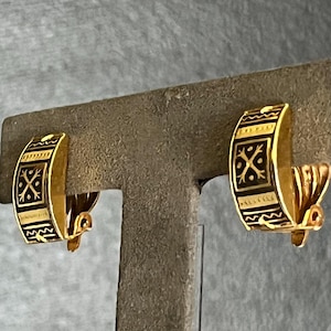 May include: A pair of gold-tone clip-on earrings featuring a curved, semi-circular design. Each earring is decorated with black enamel accents, including a central square motif with a cross pattern, flanked by horizontal bands of geometric lines.