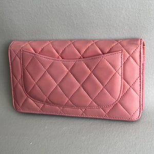 May include: A pink quilted leather wallet with a diamond pattern. The wallet has a flap closure and a pocket on the back. The stitching is visible and matches the color of the leather. The wallet is rectangular.