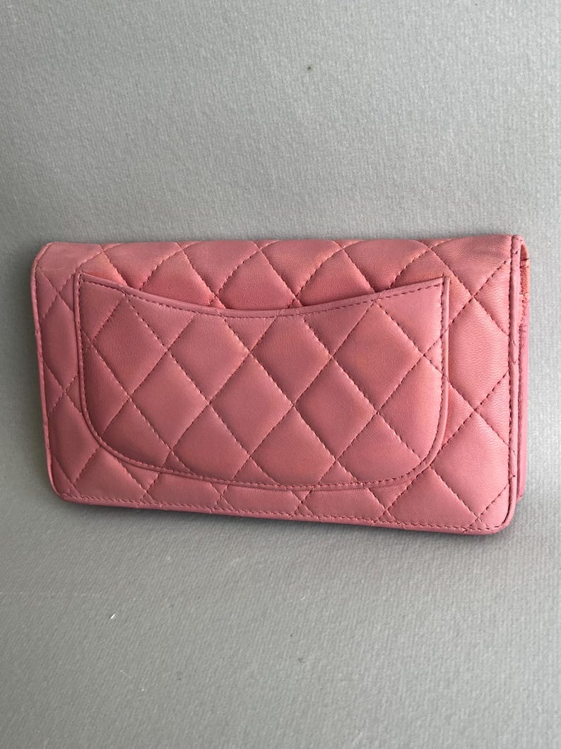 May include: A pink quilted leather wallet with a diamond pattern. The wallet has a flap closure and a pocket on the back. The stitching is visible and matches the color of the leather. The wallet is rectangular.
