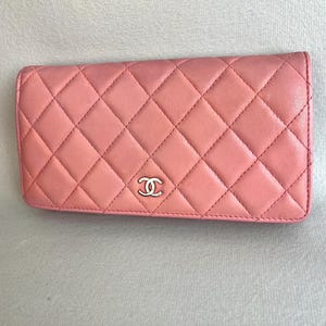 May include: A pink, quilted leather wallet with a silver-tone logo. The wallet features a diamond pattern and a rectangular shape. The stitching is visible, and the wallet appears to be made of high-quality materials.