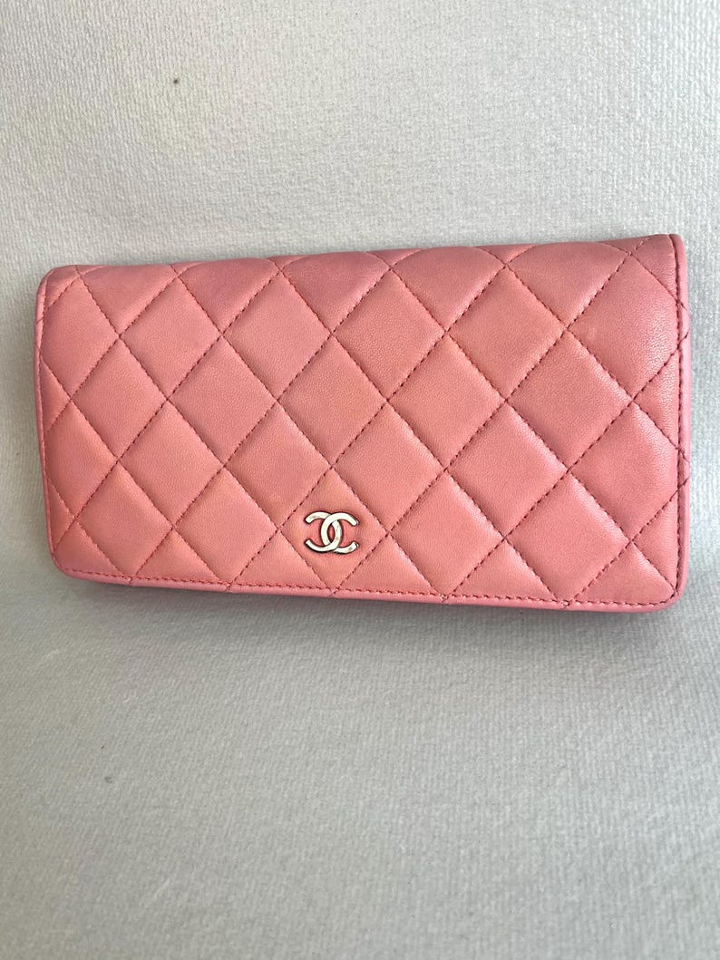May include: A pink, quilted leather wallet with a silver-tone logo. The wallet features a diamond pattern and a rectangular shape. The stitching is visible, and the wallet appears to be made of high-quality materials.