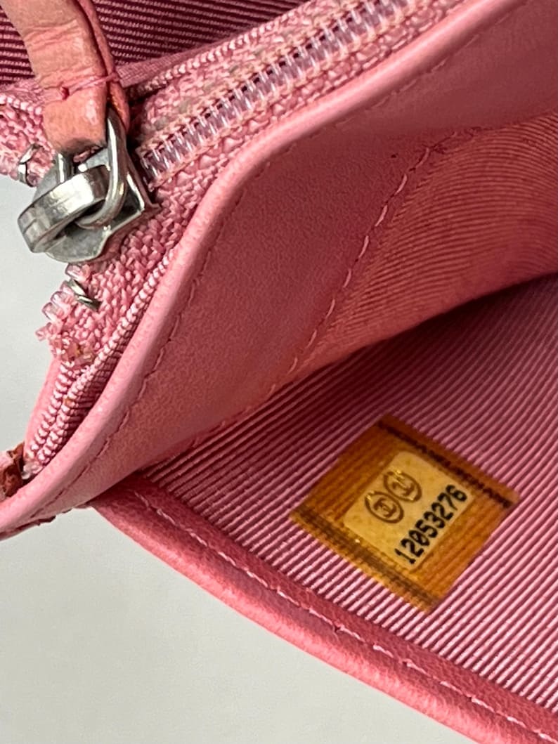 May include: Close-up of a pink leather wallet, partially open, revealing a light pink interior lining. A silver zipper and a small, rectangular label with the number "12053276" are visible. The wallet appears to be made of high-quality materials.