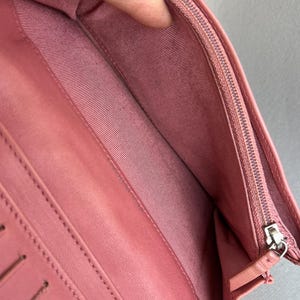 May include: Open pink leather wallet with multiple card slots and a zippered compartment. The interior features a pink fabric lining. The wallet is open, revealing the inside compartments and stitching details.