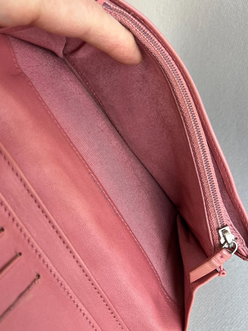 May include: Open pink leather wallet with multiple card slots and a zippered compartment. The interior features a pink fabric lining. The wallet is open, revealing the inside compartments and stitching details.