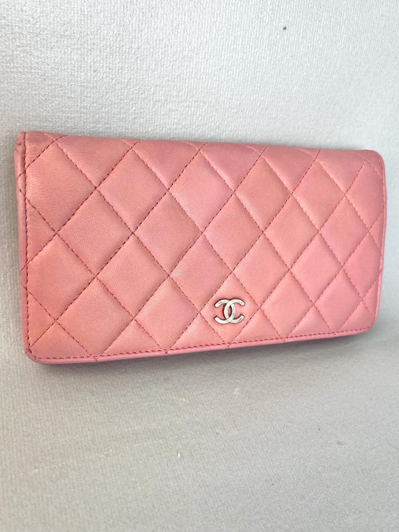 May include: A pink quilted leather wallet with a silver-tone logo. The wallet has a diamond pattern and a rectangular shape. The stitching is visible, and the wallet appears to be made of high-quality materials.