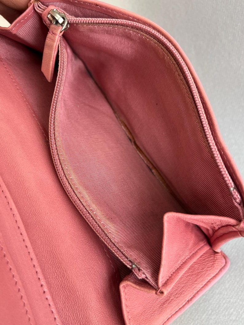 May include: Open pink leather wallet with a silver zipper and a pink fabric interior. The wallet is open, revealing the inside compartments and a coin pouch. The leather appears to be smooth and the wallet is empty.