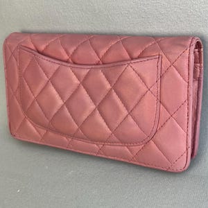 May include: A dusty rose-colored, quilted leather wallet. The wallet features a diamond pattern and a small pocket on the back. The edges are rounded, and the wallet appears to be closed.