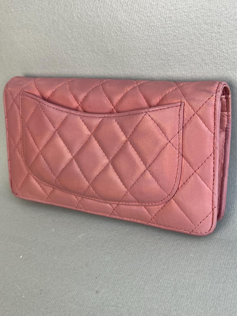 May include: A dusty rose-colored, quilted leather wallet. The wallet features a diamond pattern and a small pocket on the back. The edges are rounded, and the wallet appears to be closed.