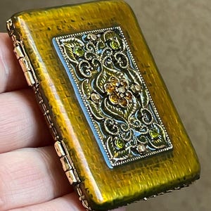 May include: A rectangular, olive-green box with a hinged lid. The box features a decorative, ornate rectangular panel with a floral design, embellished with green and amber-colored gemstones. The box has a metallic, gold-colored trim.