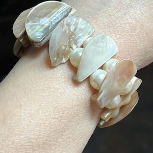 May include: A bracelet featuring iridescent, fan-shaped shell pieces and round, off-white beads. The bracelet is a mix of pearlescent and neutral tones, creating a natural and elegant look. The bracelet is worn on a wrist.