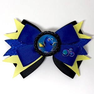 Peut inclure: Un nœud bleu et jaune avec un centre noir représentant un personnage du film Le Monde de Dory. Le personnage est un poisson chirurgien bleu avec une nageoire jaune. Le nœud a un ruban bleu pailleté et un ruban noir avec un ruban jaune sur le dessus.
