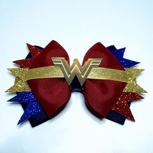 Puede incluir: Un lazo para el pelo con temática de Wonder Woman. Presenta un lazo burdeos con una banda dorada con purpurina y un emblema "W" de metal dorado. Decorado con estrellas doradas, rojas y azules con purpurina.