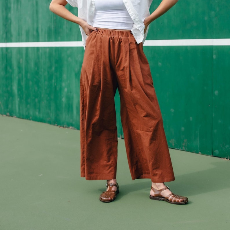 May include: A person wearing a white shirt and brown wide-leg pants with a loose fit. The pants have a wide waistband and are made of a soft, flowing fabric. The person is wearing brown sandals.