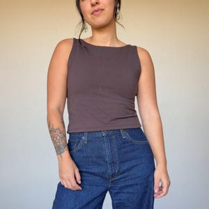 May include: A sleeveless, dark brown vest top with a scoop neckline. The top is paired with dark blue denim jeans. The model is wearing earrings and has a tattoo on their arm. The background is a solid, neutral colour.