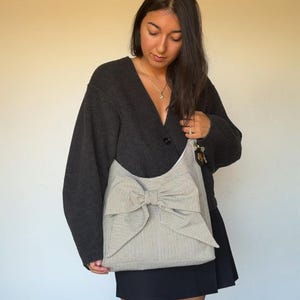 Op de afbeelding: Een grote tote bag met een opvallend strikdesign. De tas heeft een crème- en grijs gestreept patroon en een grote strik in het midden. De persoon draagt een donkergrijs vest en een zwarte rok.