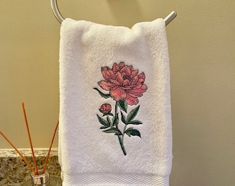 May include: White hand towel with a detailed pink peony floral design. The towel is hanging on a silver towel ring. A glass diffuser with reeds sits on the counter below.