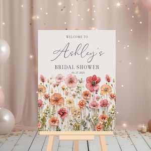 May include: A white welcome sign with a floral design and the text "Welcome to Ashley's Bridal Shower 03.27.2025".