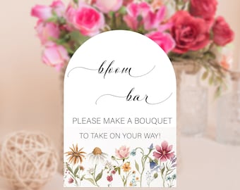 May include: A white sign with an arch shape at the top, with the text "bloom bar" in cursive script. The sign also says "Please make a bouquet to take on your way!" and features a watercolor illustration of a floral bouquet at the bottom.