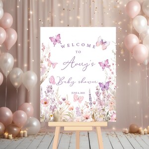 May include: Welcome to Avery's Baby Shower sign with a floral design featuring butterflies and lavender. The sign is printed on white cardstock and has a purple and pink color scheme. The date of the shower is June 6, 2025.