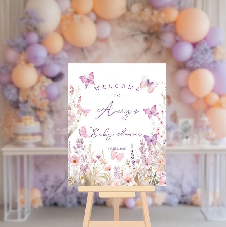May include: Welcome to Avery's Baby Shower sign with a purple floral and butterfly design. The sign is on a wooden easel and says "Welcome to Avery's Baby Shower" with the date June 6, 2023.