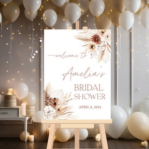 May include: A white welcome sign with a brown easel, featuring a floral design with dried pampas grass and the text "Welcome to Amelia's Bridal Shower April 8, 2024".