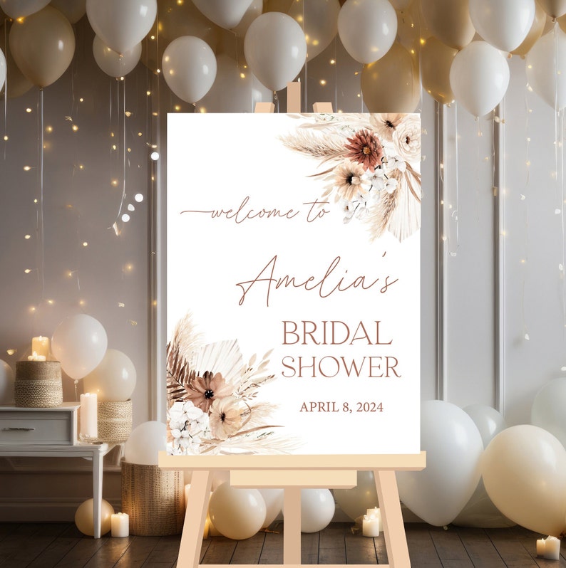 May include: A white welcome sign with a brown easel, featuring a floral design with dried pampas grass and the text "Welcome to Amelia's Bridal Shower April 8, 2024".