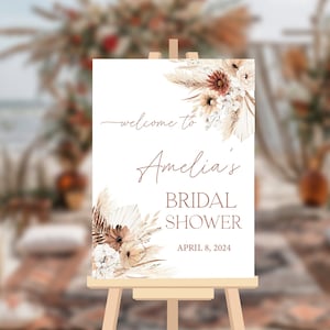May include: A white welcome sign with brown text that reads "Welcome to Amelia's Bridal Shower April 8, 2024". The sign is decorated with a watercolor illustration of dried pampas grass and flowers in shades of brown, white, and beige.