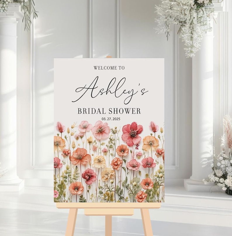 May include: A white welcome sign with a floral border and the text "Welcome to Ashley's Bridal Shower 03.27.2025".