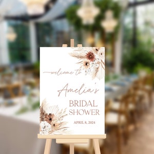 May include: A white welcome sign with a floral design in shades of brown and white. The sign reads "Welcome to Amelia's Bridal Shower April 8, 2024".
