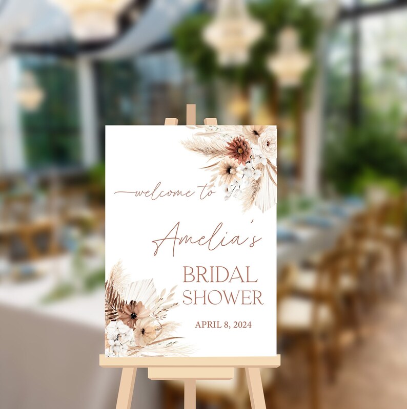 May include: A white welcome sign with a floral design in shades of brown and white. The sign reads "Welcome to Amelia's Bridal Shower April 8, 2024".