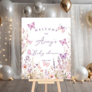 May include: Welcome to Avery's Baby Shower sign with a purple and white floral design featuring butterflies. The sign is on a wooden easel and has the date June 6, 2025.
