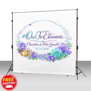 May include: A white backdrop with a floral design featuring the text "#Oui To Etienne", the names "Chandria & Fritz Gerald", and the date "05.23.2026". The design includes gold wedding rings and purple and teal flowers.