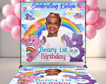 May include: A birthday party backdrop and floor mat featuring a photo of a child, Care Bears, a rainbow, and the text "Celebrating Kaliya" and "Beary 1st Birthday." Pink balloons are on either side.