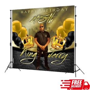 May include: A personalized birthday backdrop featuring a man in front of a cityscape, with gold and black balloons, crowns, and the text "Happy Birthday 45th King Jerry". The backdrop is supported by a black stand.