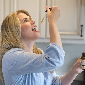 May include: A woman in a light blue shirt takes a dropper full of liquid from a small amber bottle labelled "Ritual"