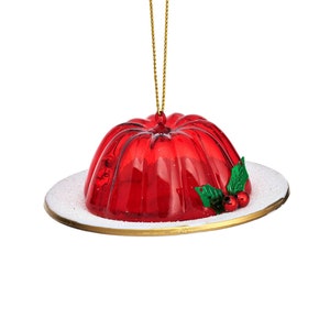 May include: Red glass Christmas ornament shaped like a bowl of red jelly with a green holly sprig and red berries on a white plate with a gold rim.
