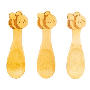 May include: Three wooden spoons with a bee design. The spoons are made of natural bamboo and have a light brown color. Each spoon has a cute cartoon bee face with a smile and closed eyes.