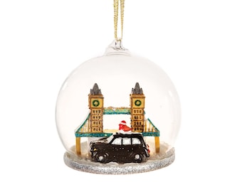May include: A glass Christmas ornament featuring a miniature model of Tower Bridge in London, England. A black taxi cab is driving across the bridge, and the ornament is decorated with silver glitter.