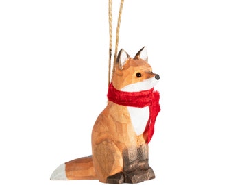 May include: A wooden fox ornament with a red scarf. The fox is sitting, with a white chest and tail tip. The ornament hangs from a natural twine string. The fox has a carved appearance.