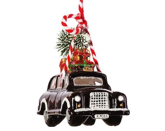 May include: A black glass Christmas ornament shaped like a vintage taxi with a red and white striped ribbon, a small green tree, and wrapped gifts on top. The taxi has a "TAXI" sign on the roof and a "XMAS" license plate.