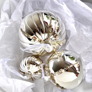 May include: Three silver Christmas ornaments with a swirl design and white and gold embellishments. The ornaments are on a white background.
