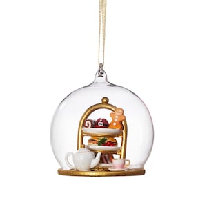 May include: A clear glass Christmas ornament shaped like a dome, featuring a miniature three-tiered serving stand with pastries, a teapot, and a teacup. The ornament has a gold-colored base and a gold hanging string.