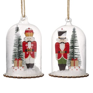 May include: Two glass dome ornaments with wooden bases. Each dome features a nutcracker figurine, a small Christmas tree, and a wrapped gift. The nutcrackers are wearing red and green uniforms with gold accents.