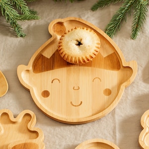 May include: A wooden elf-shaped plate with a smiling face and a small pastry on top. The plate is made of natural bamboo and has a light brown color.