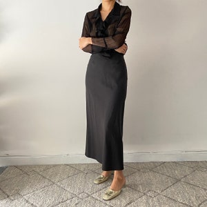 May include: A black sheer blouse with a ruffled neckline and long sleeves paired with a black maxi skirt. The model is wearing gold sandals.