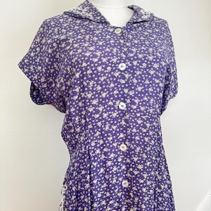 May include: A purple short-sleeved dress featuring a small white floral pattern, a collared neckline with white embroidered details, and a front button-down closure with round white buttons. The garment is displayed on a mannequin torso.