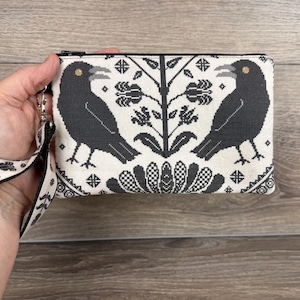 May include: A rectangular zippered pouch featuring a black and white cross-stitch pattern of two crows facing a central floral motif. The pouch includes a matching wrist strap with a repeating black floral design on a white background.