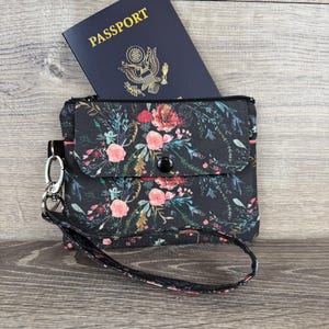 May include: A black floral passport wallet with a wrist strap, holding a blue passport. The wallet features a button closure and a silver-tone clasp. The passport is visible with the word "PASSPORT" in gold lettering.